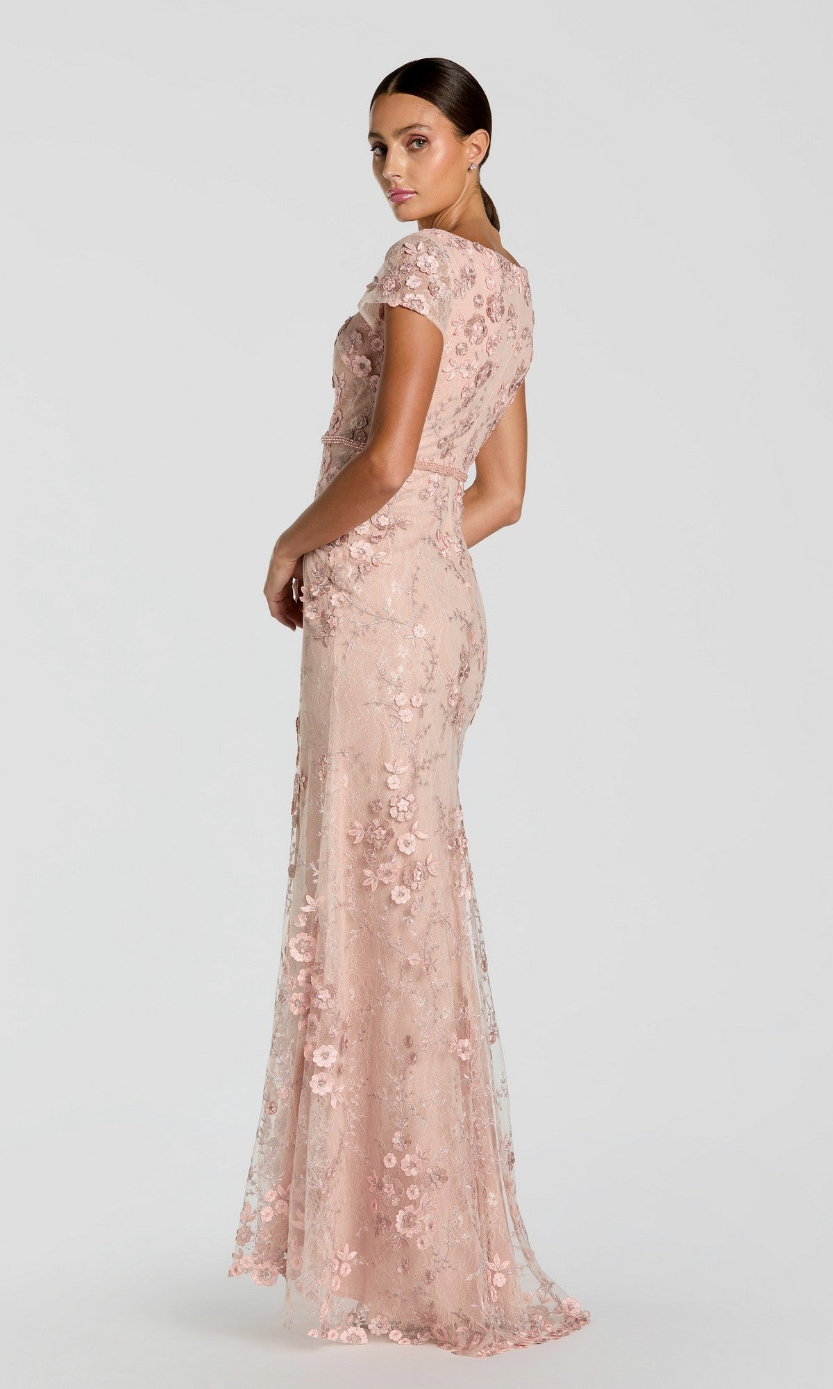 A woman stands slightly turned away, wearing the Long Formal Dress 67711 by Mac Duggal— a light pink, floor-length fitted gown with floral embroidery—against a plain light background.